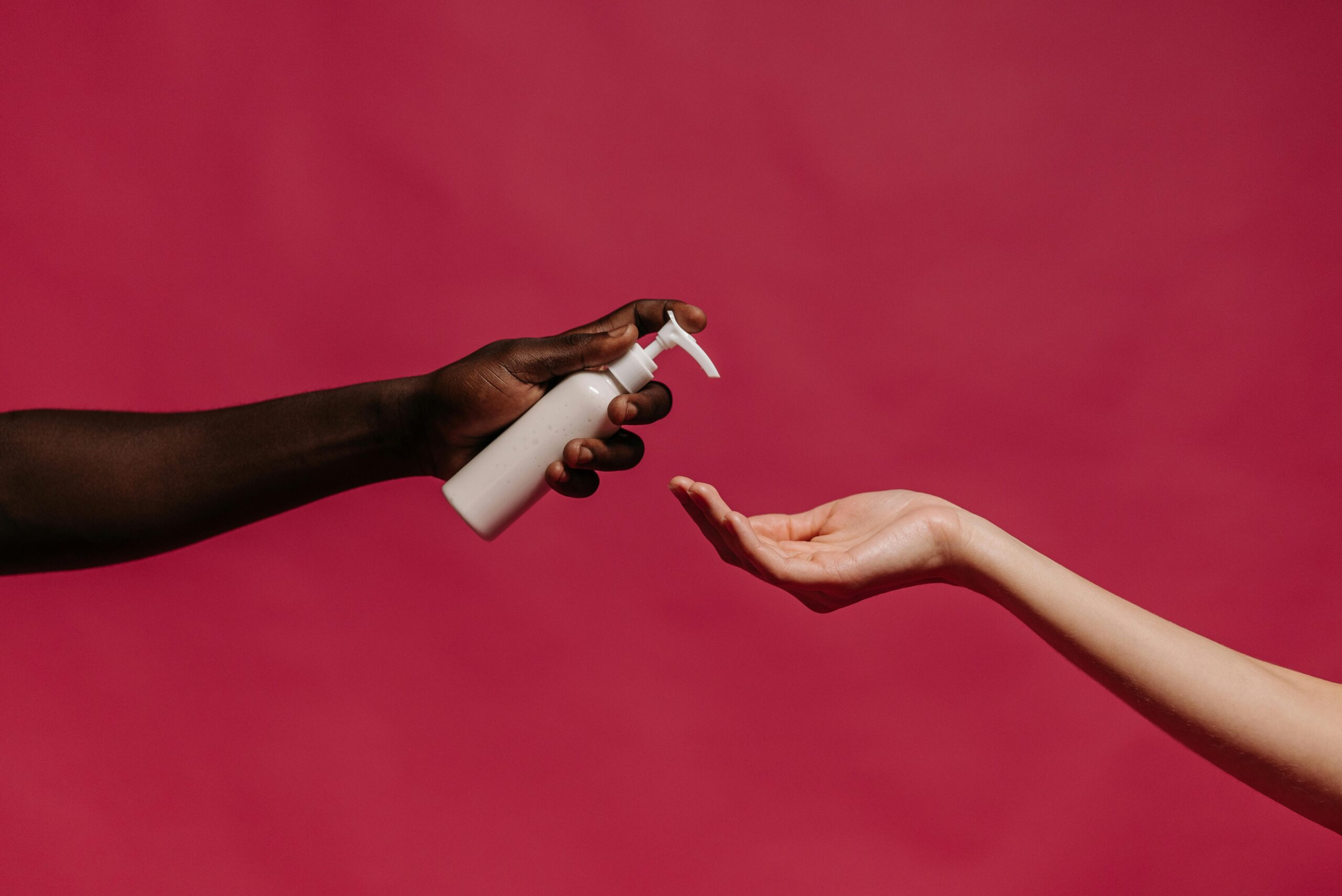 Close-up of two hands interacting with a lotion bottle on a vibrant pink background, symbolizing diversity and skincare.